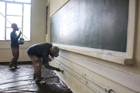 Sala de aula é preparada para pintura. Um trabalhador está raspando um pedaço de parede. Outro, ao fundo, segura uma lata de tinta e o cabo de um rolo de pintura. #Paratodosverem