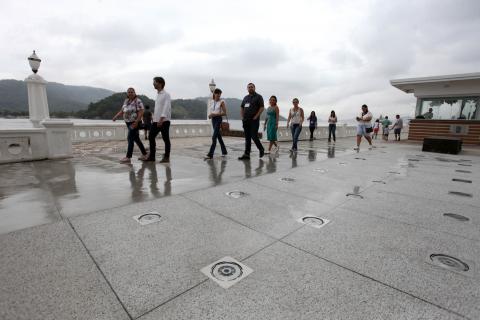 Pessoas percorrem calçadão de avenida. Ao lado direito da imagem aparecem os jatos de fonte interativa. #Pracegover