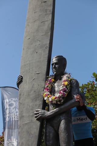 Estátua de pioneiro do surfe, com a respectiva prancha, está com um colar de flores. #Pracegover