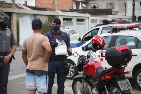 #pracegover Mascarados, policial militar e guarda municipal conversam com motocilicta que está sem máscara ao lado da moto em via pública