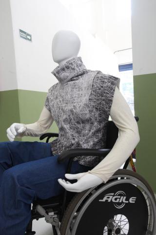 Manequim sentado em cadeira de rodas vestido com roupas específicas para sua necessidade. #Paratodosverem