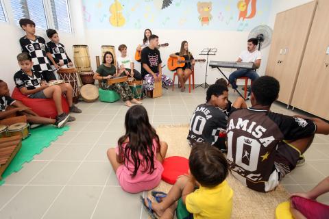 Crianças estão em uma sala de atividades. Alguns estão com instrumentos musicais. Eles estão sentados em pufs e tapetes. #Pracegover