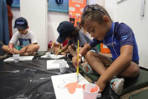 Crianças estão sentadas no chão segurando pinceis e pintando sobre folhas de papel. #pracegover