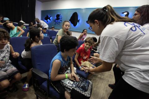 professora mostra objeto a aluno no auditório #pracegover 