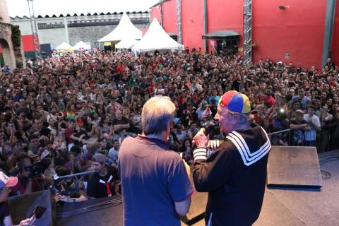 Kiko está no palco com um homem. O público lota o local. #Pracegover