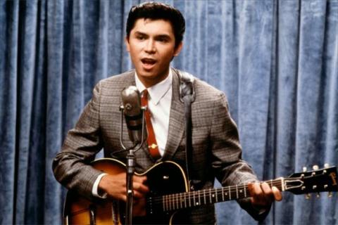 Personagem de La Bamba toca guitarra diante de um microfone. #Pracegover