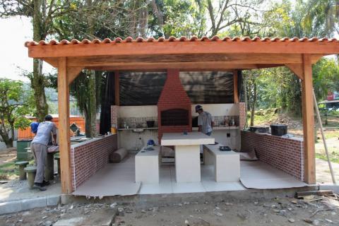 Quiosque coberto, com mesa e bancos de alvenaria no centro e churrasqueira ao fundo. #Paratodosverem