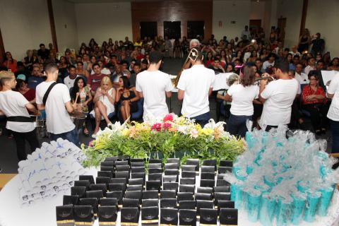 O auditório visto da plateia para o público. Em primeiro plano há uma mesa com os canudos dos formandos e outras peças. À frente da mesa, músicos de uma banda estão se apresentando. #Pracegover