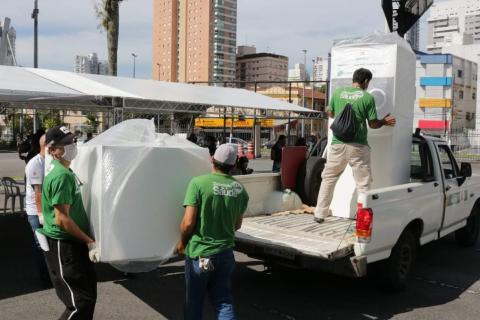Homens retiram grandes caixas de caçamba de veículo. Atrás deles há uma tenda. #Paratodosverem