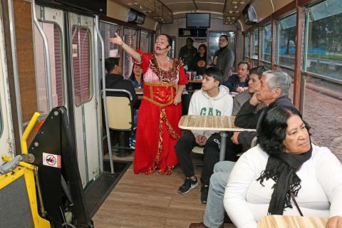 Teodora, a senhora do café, vestida com um longo vermelho, fala aos passageiros do bonde. #Pracegover