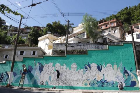 O grande muro ao fundo, com casas ao fundo,. Mulheres estão grafitando a obra. #Paratodosverem