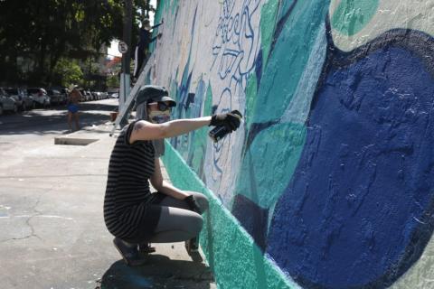 Mulher agachada pinta com grafite a arte desenhada em muro. #Paratodosverem