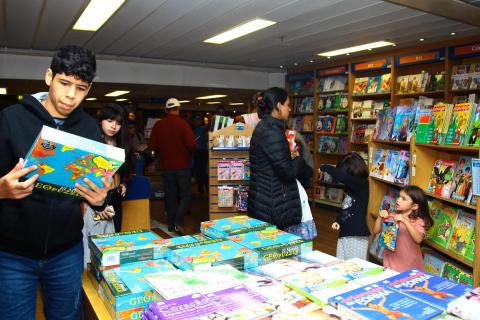 Jovem, em primeiro plano, segura livro. Ele está ao lado de uma mesa cheia de publicações. Há adultos e crianças e no ambiente. #Pracegover