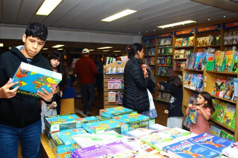 Jovem, em primeiro plano, segura livro. Ele está ao lado de uma mesa cheia de publicações. Há adultos e crianças e no ambiente. #Pracegover