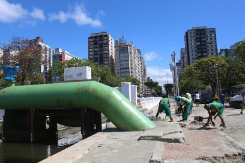 Homens fazem manutenção de mosaico em calçada ao lado de canal. #Paratodosverem