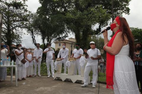 Mulher canta com homens ao fundo tocando instrumentos de percussão. 