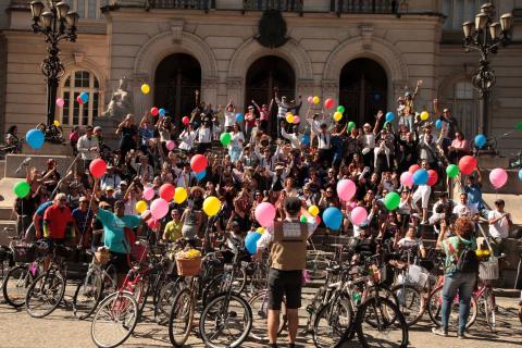 Ciclistas estão na Praça Mauá. O volume é grande. Eles ocupam as escadarias da Prefeitura com suas bicicletas e balões de gás coloridos. #Pracegover