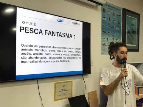 Homem fala ao microfone. Ao fundo há uma tela onde se lê Pesca Fantasma. Um texto segue abaixo. #Paratodosverem