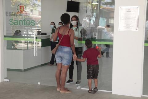 Mulher segurando a mão de criança está na porta da unidade conversando com uma recepcionista. #Paratodosverem