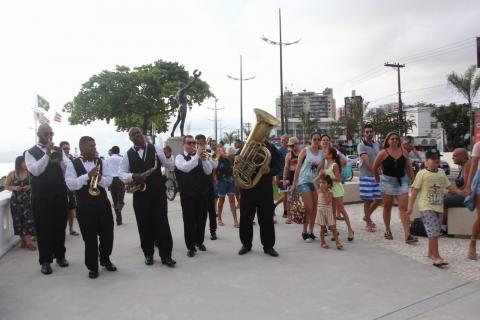 Conjunto de sopro se apresenta no calçadão. Eles estão caminhando. Público acompanha ao lado. #Pracegover