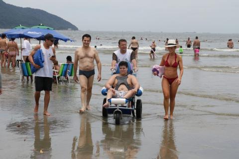 Pessoa com deficiência passeia em cadeira anfíbia na beira do mar ao lado de familiares e monitores. #pracegover