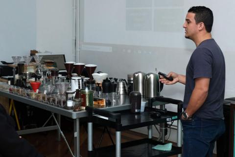 Treinamento sobre café. Várias jarras, bules e filtros estão sobre uma mesa. Um homem está falando. #Pracegover