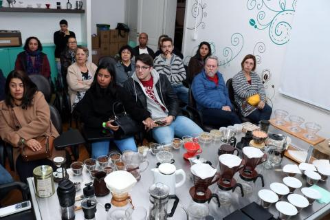 Público está sentado em sala diante de mesa cheia de jarras, filtros e potes com exemplares de grãos de café. #Pracegover