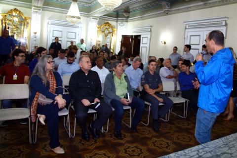 Apresentação de projeto no paço municipal. Público está sentado atendo às explicações feitas por um homem. #Pracegover