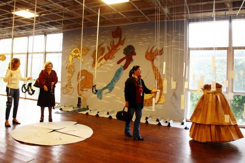 Sala de exposição com linhas com objetos pendurados na ponta. Ao fundo há um painel com sereias e uma instalação de madeira. #Pracegover