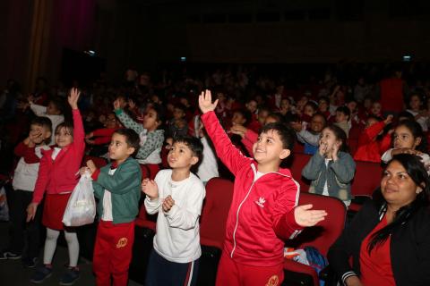 crianças na plateia levantam as mãos #pracegover 