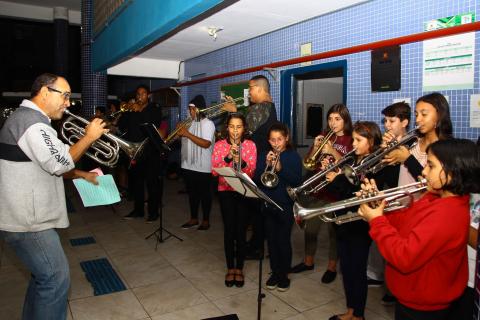 Estudantes estão em pé em um salão tocando instrumentos de sopro. À frente deles há um professor que também está tocando. #Pracegover