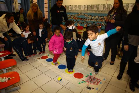 Crianças participam de atividade na sala. Elas estão pulando sobre círculos marcados no chão da sala. #Pracegover