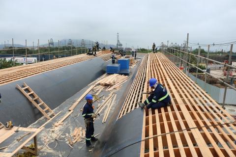Vista geral da estrutura, com madeira aparente e homens trabalhando no topo. #Pracegover