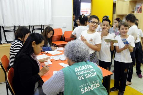 agentes trabalham na mesa enquanto crianças fazem fila para vacinação #pracegover 