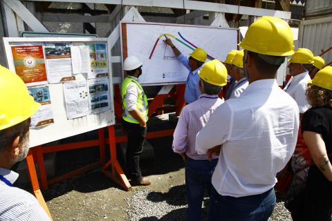 Projeto fixado em painel ao fundo é mostrado a homens e mulheres com capacetes na cabeça. #Pracegover