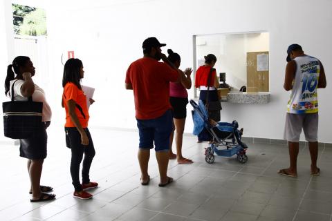 Homens e mulheres estão em fila em frente a guichê fechado por vidro transparente. Há também um carrinho de bebê no ambiente. #Pracegover