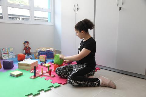 Sala de brinquedos. Menina está sentada no chão sobre tapete manuseando peças. #Paratodosverem