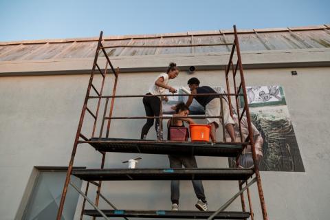 Painéis com imagens são afixados em parede. Jovens fazem esse trabalho em cima de andaimes. #Paratodosverem