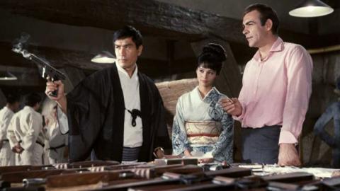 Um casal de japoneses ao lado de James Bond. O japonês está segurando uma arma e a mulher está vestida de gueixa. #Pracegover