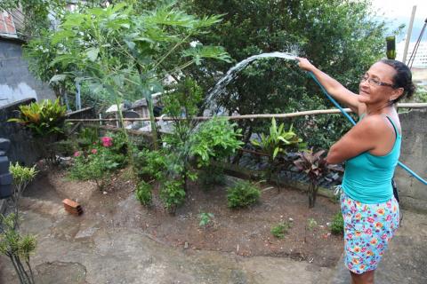 Socorro segura mangueira e rega plantas na frente de sua casa. #Paratodosverem