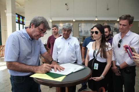 Assinatura do convênio. Homem está assinando documento sobre uma mesa. Ele está de pé ao lado de outras pessoas que assistem. #Pracegover