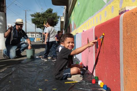 Crianças pintam parte de baixo do muro. Uma criança em primeiro plano segura um pincel. #Pracegover