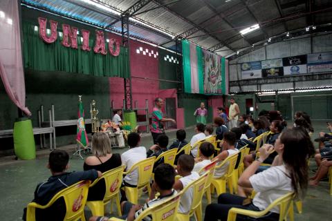 Estudantes estão sentados em cadeiras na quadra da escola de samba. Eles estão atentos a um homem que fala ao gruupo. #Paratodosverem