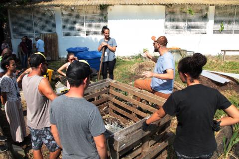 Várias pessoas em torno de uma composteira ouvindo orientações de um homem. #Paratodosverem