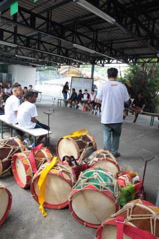 Instrumentos de percussão estão o chão. Há pessoas em torno. #Pracegover