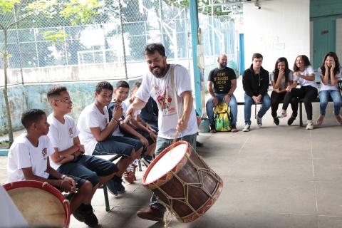 Orientador toca instrumento de percussão diante de alunos que estão sentados. #Pracegover