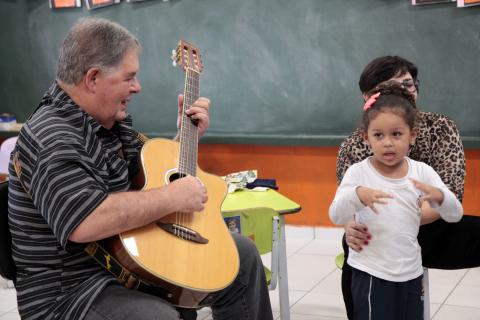 Vovô toca violão com criança ao seu lado. A menina mexe as mãos. #Pracegover