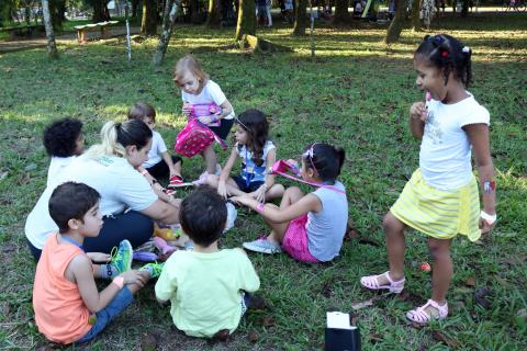 Crianças estão sentadas na grama e brincam sob orientação de uma mulher. Uma menina está em pé #Pracegover