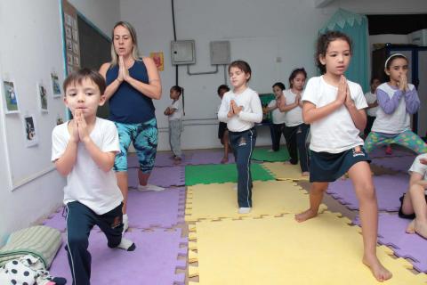 Crianças e professora fazem movimento. Elas estão em pé, com um dos pés avançados para a frente e flexionados. Todas estão como as mãos unidas na frente do corpo. #Pracegover