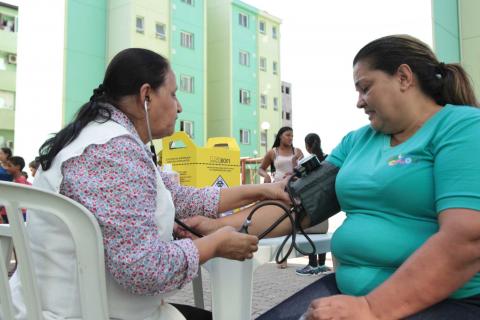 Mulher mede a pressão arterial de outra mulher. Elas estão sentadas, com uma mesa entre as duas. #Pracegover